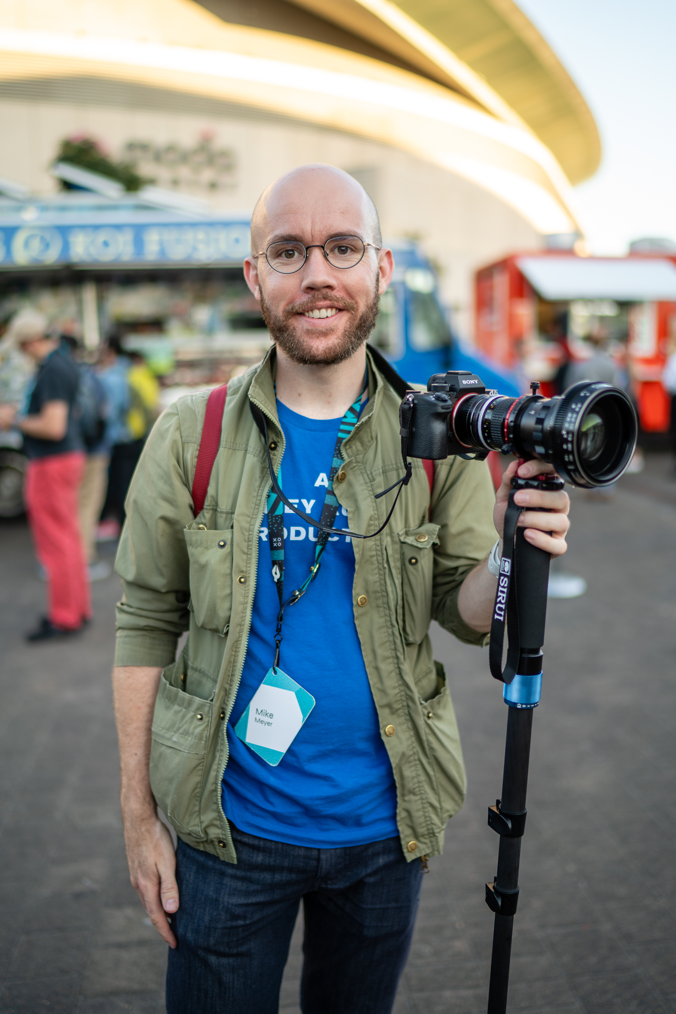 Portrait of me, a bald guy in a blue t-shirt and a green jacket. I’m standing and smiling at the camera. In my left hand I’m holding a monopod like a staff. Atop the monopod is a Sony mirrorless camera with a comically large anamorphic lens.